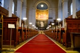 An interior of a grand church with a symmetrical view down the central aisle. The aisle is carpeted in red, flanked by ornate wooden pews. At the far end, a large statue stands beneath a vibrant mural depicting religious figures, with a domed ceiling above adorned with a star design. Tall columns line the sides, supporting the arched roof, and soft lighting from lamps illuminates the space.