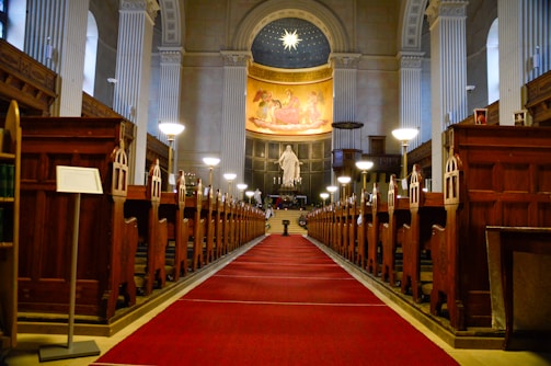An interior of a grand church with a symmetrical view down the central aisle. The aisle is carpeted in red, flanked by ornate wooden pews. At the far end, a large statue stands beneath a vibrant mural depicting religious figures, with a domed ceiling above adorned with a star design. Tall columns line the sides, supporting the arched roof, and soft lighting from lamps illuminates the space.