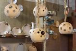 Several ceramic lamps with spherical, perforated designs hang from ropes. In the background, there are shelves displaying various plates and dishes with floral patterns. The setting appears to be a cozy, handcrafted decor store or pottery shop.