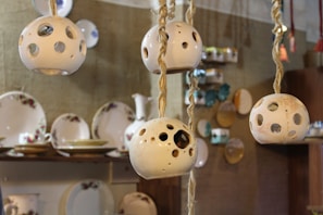 Several ceramic lamps with spherical, perforated designs hang from ropes. In the background, there are shelves displaying various plates and dishes with floral patterns. The setting appears to be a cozy, handcrafted decor store or pottery shop.