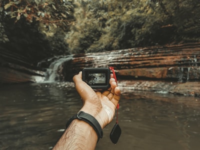 A person holds a small camera in their hand, capturing an image of a serene waterfall cascading over layered rocks surrounded by lush green foliage. The camera's display shows a smaller version of the waterfall scene. The image is taken at an angle where the arm is extended towards the waterfall, with water visible in the foreground.
