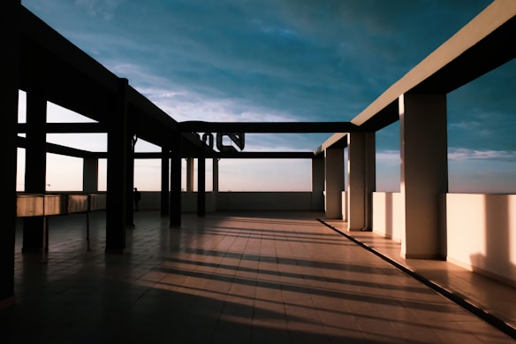 A rooftop terrace with a minimalist architectural design features several large, rectangular pillars casting long shadows across the tile floor. The sky, filled with clouds, creates a dramatic backdrop with gradients of blue and hints of orange from the sunset.