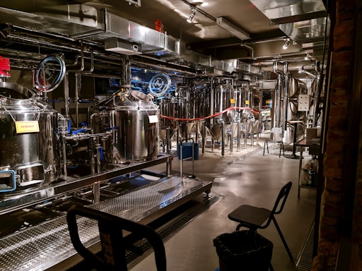A brewery with stainless steel tanks and machinery, complete with pipes and gauges for brewing operations. The setting includes various hoses, control panels, and some chairs. Industrial lighting illuminates the workspace, highlighting the modern, industrial aesthetic.