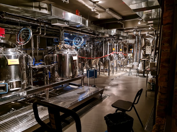 A brewery with stainless steel tanks and machinery, complete with pipes and gauges for brewing operations. The setting includes various hoses, control panels, and some chairs. Industrial lighting illuminates the workspace, highlighting the modern, industrial aesthetic.