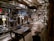 A brewery with stainless steel tanks and machinery, complete with pipes and gauges for brewing operations. The setting includes various hoses, control panels, and some chairs. Industrial lighting illuminates the workspace, highlighting the modern, industrial aesthetic.