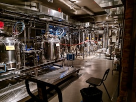 A brewery with stainless steel tanks and machinery, complete with pipes and gauges for brewing operations. The setting includes various hoses, control panels, and some chairs. Industrial lighting illuminates the workspace, highlighting the modern, industrial aesthetic.