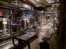 A brewery with stainless steel tanks and machinery, complete with pipes and gauges for brewing operations. The setting includes various hoses, control panels, and some chairs. Industrial lighting illuminates the workspace, highlighting the modern, industrial aesthetic.