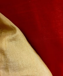 A close-up of textured fabric with a smooth, shiny golden cloth lying over a rich, vibrant red background. The fabric appears to be draped, showing natural folds and shadows, which highlight the different textures and surface qualities of the materials.