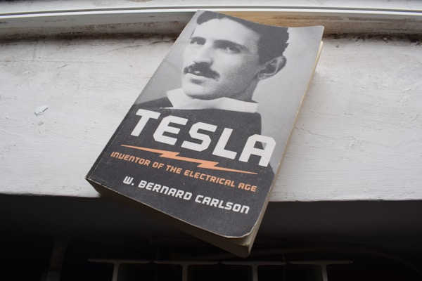 A book titled 'Tesla: Inventor of the Electrical Age' by W. Bernard Carlson is placed on a white surface. The cover features a black and white photograph of a man with a mustache, likely representing Nikola Tesla.