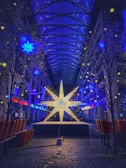 A large illuminated star sculpture stands in the center of an arcade with a glass ceiling. Blue and yellow star-shaped lights hang from the ceiling and adorn the surrounding trees, creating a festive atmosphere. The surrounding area is lined with empty benches and small trees, with leaves scattered on the ground.