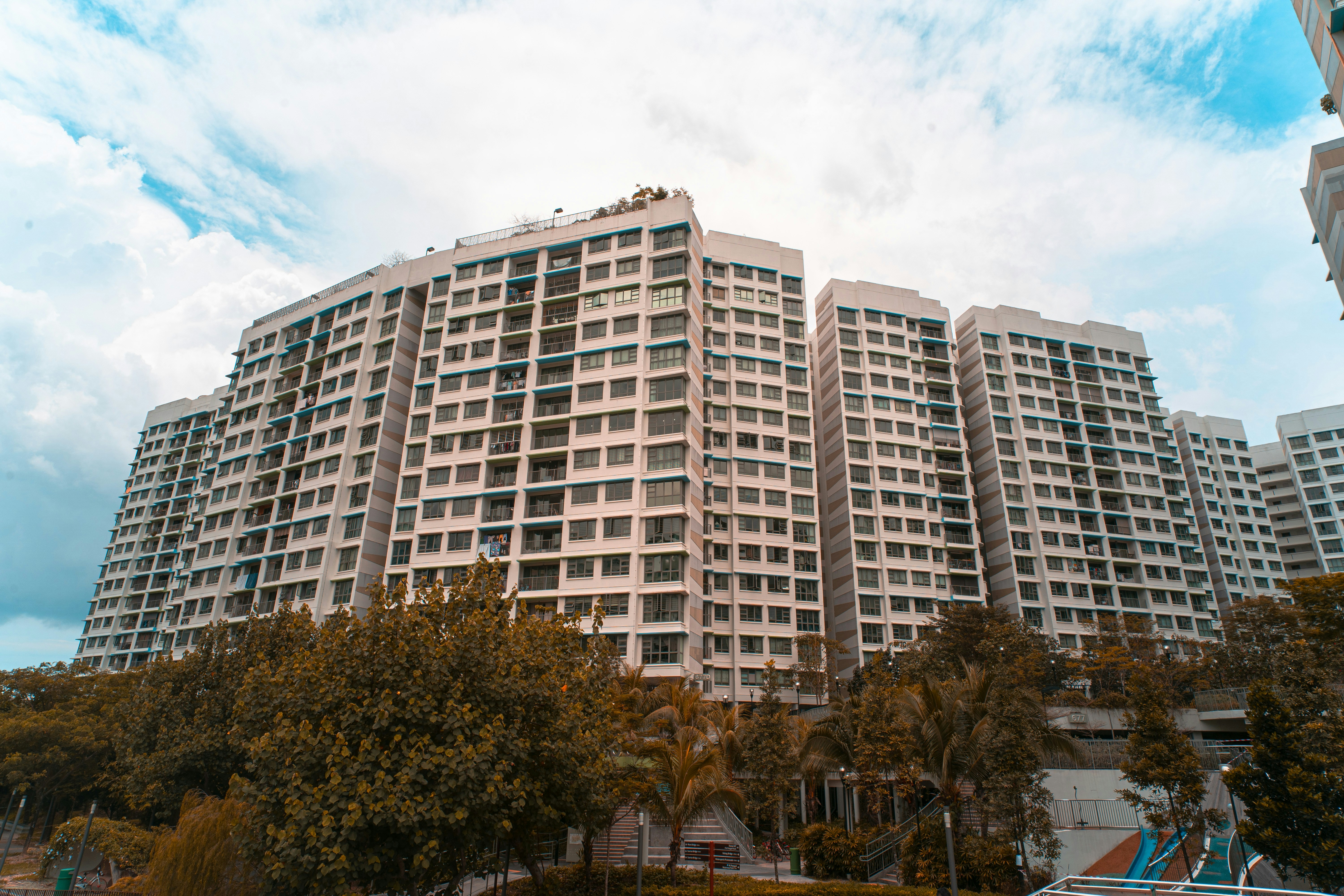 A large residential complex with multiple high-rise buildings is surrounded by greenery. The buildings have a modern architectural design with numerous windows and balconies. The sky is partly cloudy, adding a dramatic atmosphere to the scene.