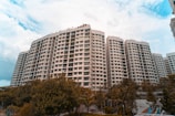 A large residential complex with multiple high-rise buildings is surrounded by greenery. The buildings have a modern architectural design with numerous windows and balconies. The sky is partly cloudy, adding a dramatic atmosphere to the scene.