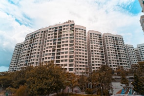A large residential complex with multiple high-rise buildings is surrounded by greenery. The buildings have a modern architectural design with numerous windows and balconies. The sky is partly cloudy, adding a dramatic atmosphere to the scene.