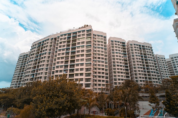 A large residential complex with multiple high-rise buildings is surrounded by greenery. The buildings have a modern architectural design with numerous windows and balconies. The sky is partly cloudy, adding a dramatic atmosphere to the scene.