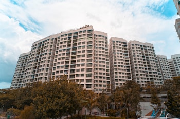 A large residential complex with multiple high-rise buildings is surrounded by greenery. The buildings have a modern architectural design with numerous windows and balconies. The sky is partly cloudy, adding a dramatic atmosphere to the scene.