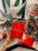 A hand holding a red gift box tied with a red and white string. In the background, a festive Christmas tree adorned with red ornaments and lights stands next to a large green houseplant. Balloons in shades of white, pink, and red rest on the floor, and a small red heart-shaped pillow is near the base of the tree.