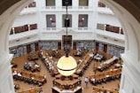 A grand, multi-story library interior with high ceilings, ornate molding, and large archways. Rows of wooden desks filled with people reading and studying are present. Bookshelves line the walls, filled with books, and large, round light fixtures hang from the ceiling.