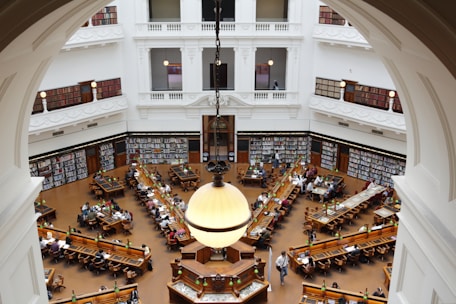 A grand, multi-story library interior with high ceilings, ornate molding, and large archways. Rows of wooden desks filled with people reading and studying are present. Bookshelves line the walls, filled with books, and large, round light fixtures hang from the ceiling.