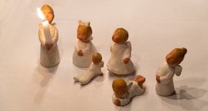 Several small, figurine-like angels are arranged on a plain surface. One angel is holding a lit candle with an expression of singing or joy. The others are in different poses, with intricate details in their white clothing and golden hair.