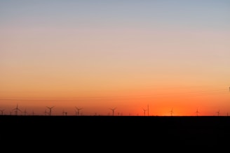 A row of wind turbines is silhouetted against a gradient sky transitioning from warm orange near the horizon to a cooler blue higher up. The setting sun creates a peaceful and serene atmosphere across the landscape with power lines faintly visible.