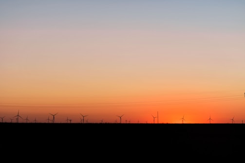 A row of wind turbines is silhouetted against a gradient sky transitioning from warm orange near the horizon to a cooler blue higher up. The setting sun creates a peaceful and serene atmosphere across the landscape with power lines faintly visible.