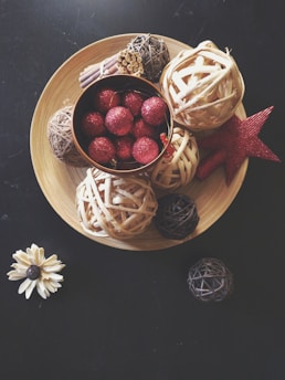 A wooden plate holds decorative woven balls, a small bunch of cinnamon sticks, a cluster of red glittery spheres, and a red woven star. Outside the plate lies a small artificial white flower and several additional woven balls, all set on a dark surface.
