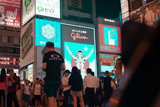 A bustling urban scene features a crowd of people walking past brightly lit billboards. One prominent sign depicts a running figure against a blue background. The area is lively with a mixture of tourists and locals, suggestive of a popular and busy district.