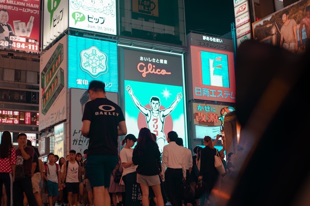 A bustling urban scene features a crowd of people walking past brightly lit billboards. One prominent sign depicts a running figure against a blue background. The area is lively with a mixture of tourists and locals, suggestive of a popular and busy district.
