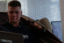 A person is sitting in a massage chair, appearing relaxed and focused. The background features a geometric textured wall and a blurred television or monitor.