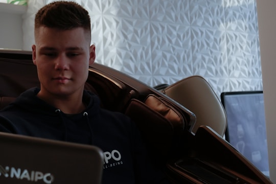 A person is sitting in a massage chair, appearing relaxed and focused. The background features a geometric textured wall and a blurred television or monitor.