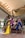 A group of six women wearing vibrant, flowing outfits stand confidently around a wooden bench in a modern indoor setting with high ceilings and large pillars. The women are posed elegantly with varied hairstyles and expressions, conveying a sense of strength and elegance.