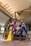 A group of six women wearing vibrant, flowing outfits stand confidently around a wooden bench in a modern indoor setting with high ceilings and large pillars. The women are posed elegantly with varied hairstyles and expressions, conveying a sense of strength and elegance.