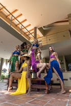 A group of six women wearing vibrant, flowing outfits stand confidently around a wooden bench in a modern indoor setting with high ceilings and large pillars. The women are posed elegantly with varied hairstyles and expressions, conveying a sense of strength and elegance.