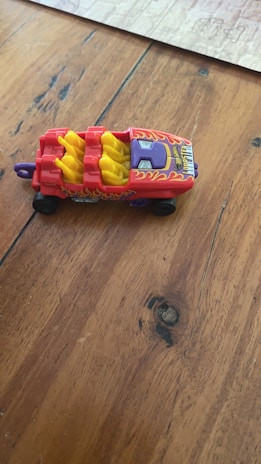 A colorful toy car with flame decals is placed on a wooden surface. The car is predominantly red with yellow seating and black wheels. It features a sleek design with hot rod styling and a label on the front.