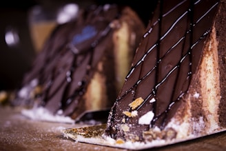 Two slices of cake are topped with a smooth layer of chocolate ganache, drizzled with thin lines of dark chocolate, and set on a wooden surface. The cake layers include textures of a soft sponge and creamy filling, sprinkled with bits of sugar and crumbs.