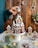 A hand sprinkles powdered sugar over a gingerbread house, set on a white pedestal. The scene includes decorated gingerbread cookies shaped like houses, stars, and trees. The background features a wooden wall adorned with additional gingerbread ornaments. Green pine branches and baubles decorate the foreground, adding to the festive atmosphere.