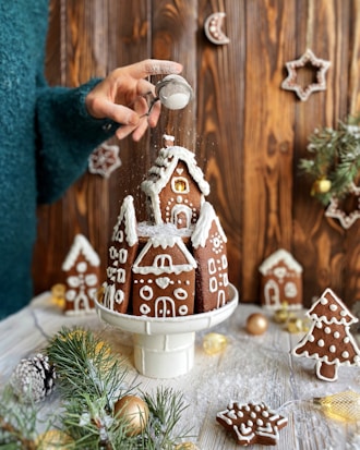 A hand sprinkles powdered sugar over a gingerbread house, set on a white pedestal. The scene includes decorated gingerbread cookies shaped like houses, stars, and trees. The background features a wooden wall adorned with additional gingerbread ornaments. Green pine branches and baubles decorate the foreground, adding to the festive atmosphere.