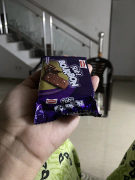A hand holding a package of Parle Fab! Bourbon biscuits, with a blurred background that includes a staircase and a black chair. The biscuit packaging is primarily purple with images of chocolate biscuits on it.