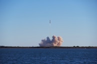 A rocket is launching into the sky from a land base near a large body of water. A plume of smoke and dust is visible at the base of the launch site, and the sky is clear with a few clouds.
