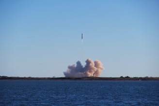 A rocket is launching into the sky from a land base near a large body of water. A plume of smoke and dust is visible at the base of the launch site, and the sky is clear with a few clouds.