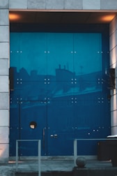 A modern, large blue glass door framed by stone tiles with reflections of buildings. The door features a circular window and minimalist handles. Warm lighting adds a cozy touch to the urban setting.