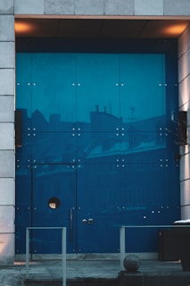 A modern, large blue glass door framed by stone tiles with reflections of buildings. The door features a circular window and minimalist handles. Warm lighting adds a cozy touch to the urban setting.