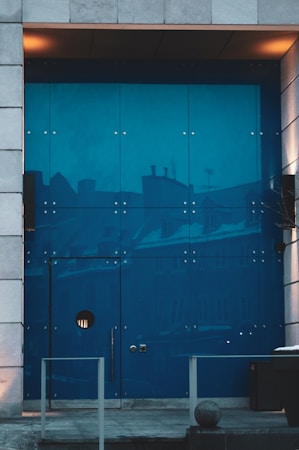 A modern, large blue glass door framed by stone tiles with reflections of buildings. The door features a circular window and minimalist handles. Warm lighting adds a cozy touch to the urban setting.