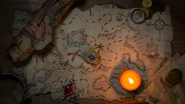 A vintage-style treasure map lies on a wooden surface, illuminated by a lit candle. The map features detailed sketches of landscapes, a ship, and a compass rose, with a red dotted line indicating a path to a marked treasure chest. Scattered around the map are antique coins, a rolled-up scroll, a ring, and decorative jewelry with a red gem.