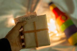 A hand holding a small, wrapped gift box with a brown twine and a thank you note attached. In the background, there is a colorful bottle-shaped decoration and blurred warm fairy lights creating a cozy atmosphere.
