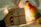 A hand holding a small, wrapped gift box with a brown twine and a thank you note attached. In the background, there is a colorful bottle-shaped decoration and blurred warm fairy lights creating a cozy atmosphere.