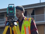 A person wearing a high-visibility safety vest and a beanie stands outdoors, operating a theodolite mounted on a tripod. The background includes the facade of a residential building with balconies.