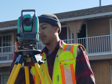 A person wearing a high-visibility safety vest and a beanie stands outdoors, operating a theodolite mounted on a tripod. The background includes the facade of a residential building with balconies.