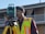 A person wearing a high-visibility safety vest and a beanie stands outdoors, operating a theodolite mounted on a tripod. The background includes the facade of a residential building with balconies.