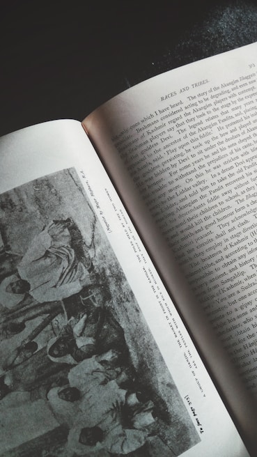 An overhead view of an open book featuring a section titled 'Races and Tribes.' The left page contains a grayscale photograph showing a group of people in traditional attire. The right page is filled with text that appears to be a narrative or historical account.